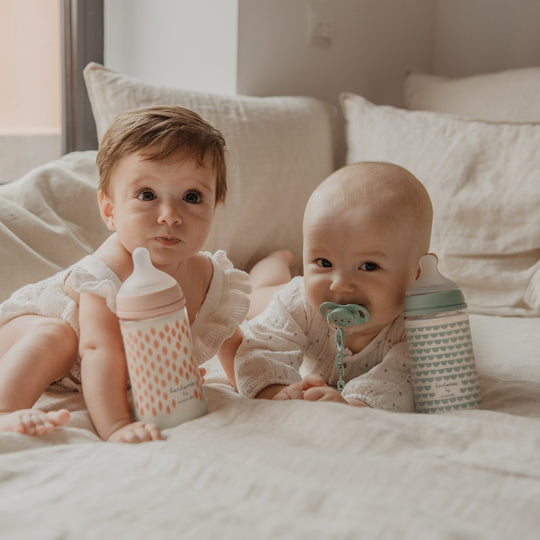 Two babies sitting on a bed with Bonhomia Glass baby bottles and a pacifier.