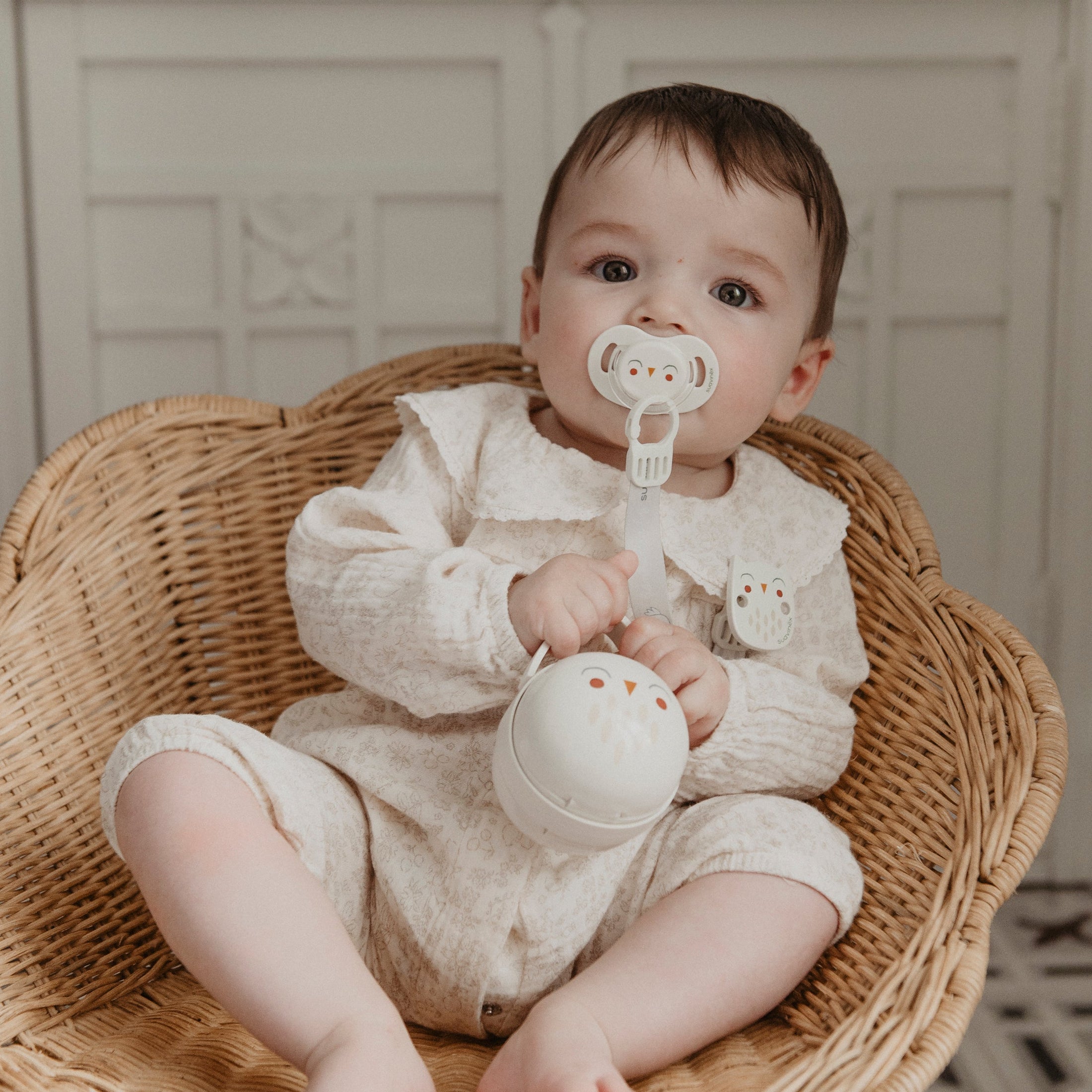 Baby sitting in a wicker chair holding a pacifier case and pacifier, with a neutral background.